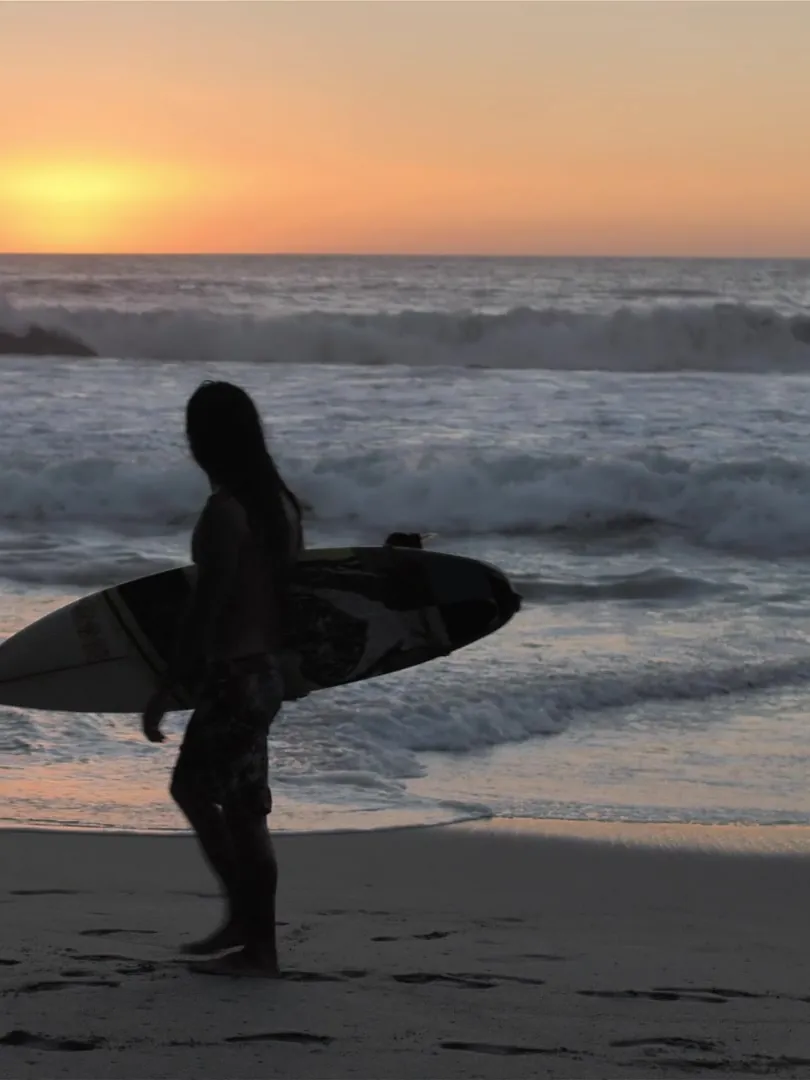 A surfer carrying a board along the water's edge at sunset