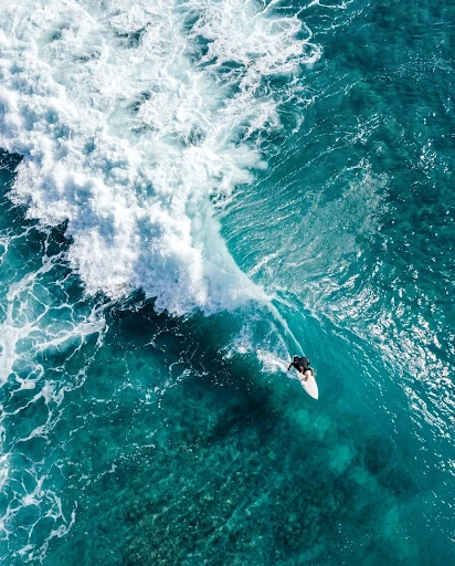 Drone view of a surfer riding a turquoise wave beside white foam