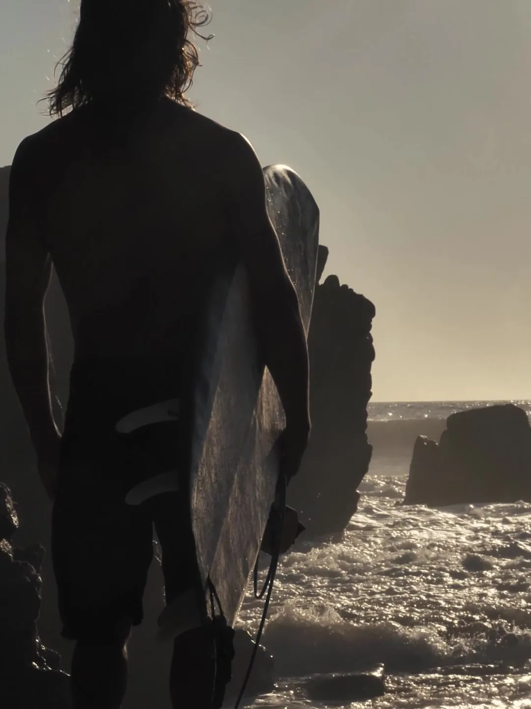 A surfer seen from behind, carrying a board towards the rocks